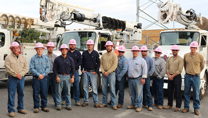 men in jeans and shirts with pink hard hats standing in front of bucket trucks