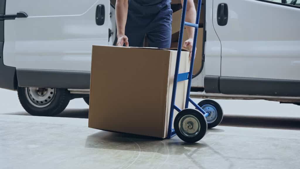 Cropped view of delivery man holding cardboard box near cart and auto outdoors