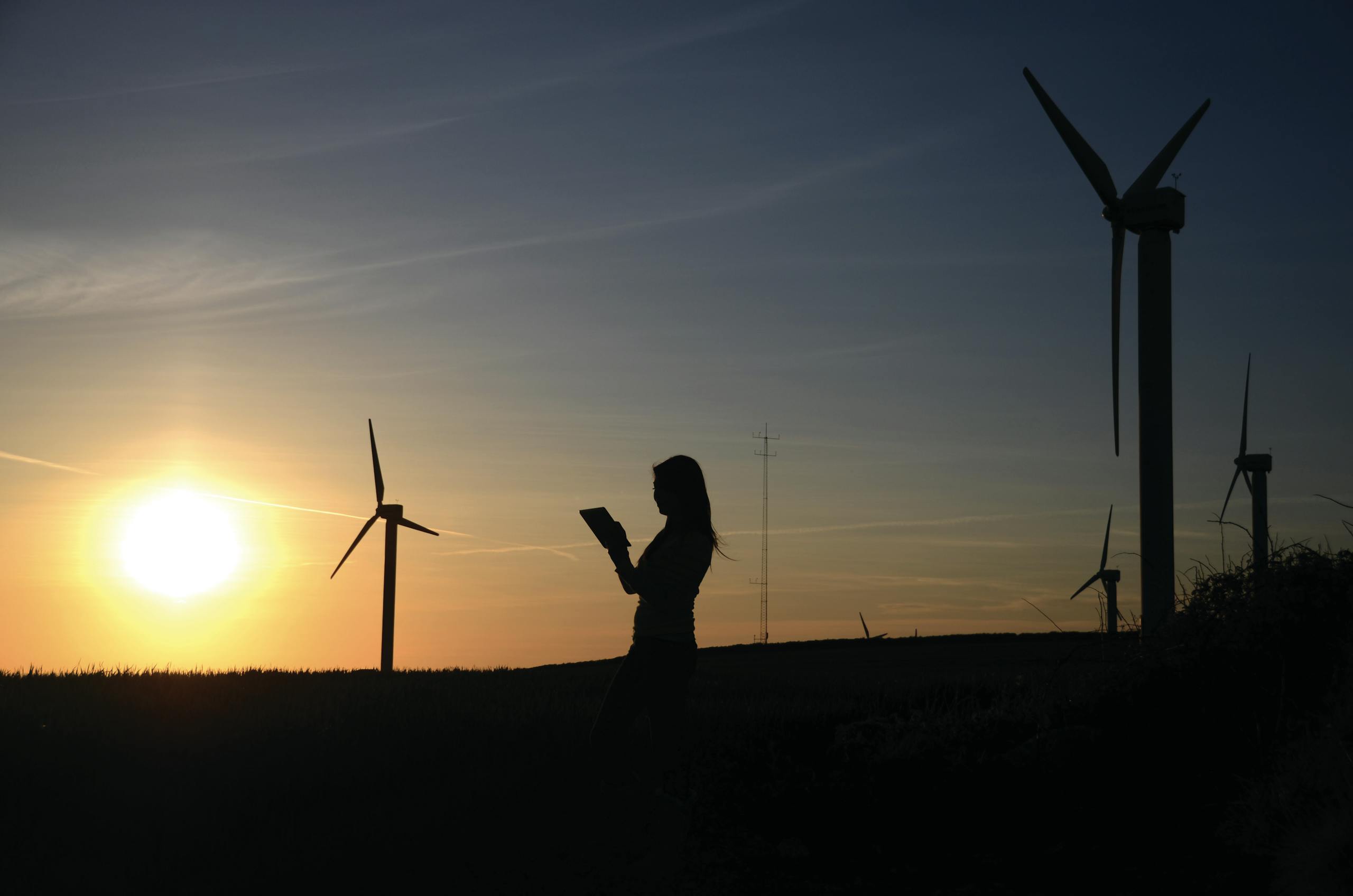 A silhouette of a woman using a device near wind turbines at sunset, representing renewable energy.