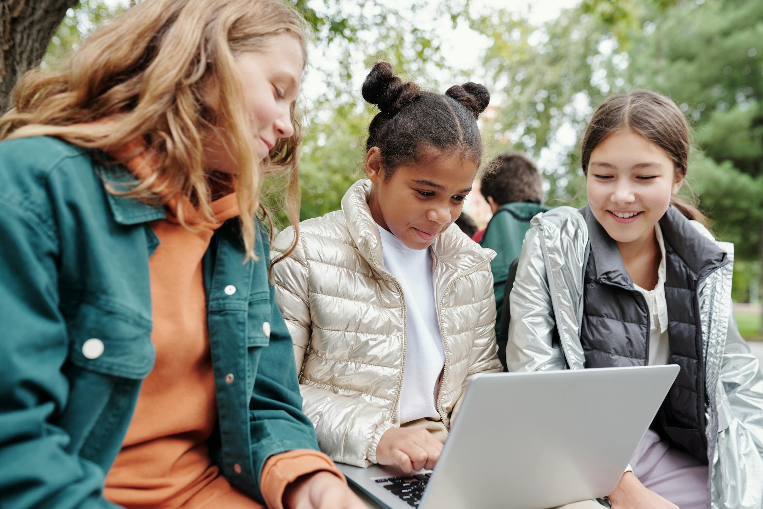 Three girls sitting together outdoors, smiling and learning on a laptop in a park setting.